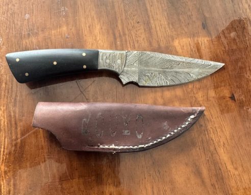 A metal knife with a black handle and a leather sheath on a wooden surface.
